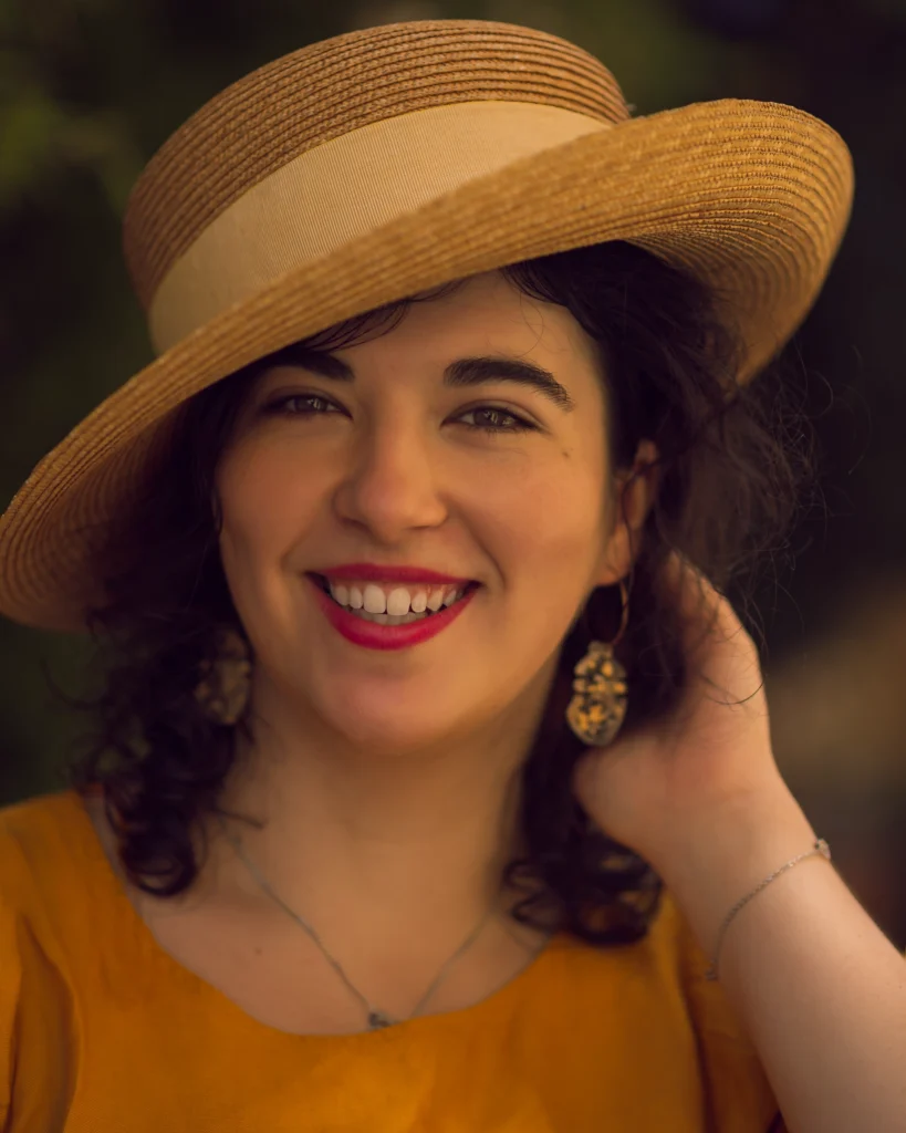 Uśmiechnięta kobieta w żółtej bluzce i kapeluszu z szerokim rondem na tle rozmytego zieleni.