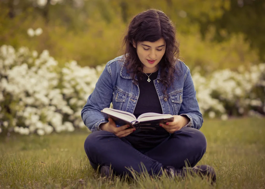 Młoda kobieta w dżinsowej kurtce czytająca książkę na trawie w parku.