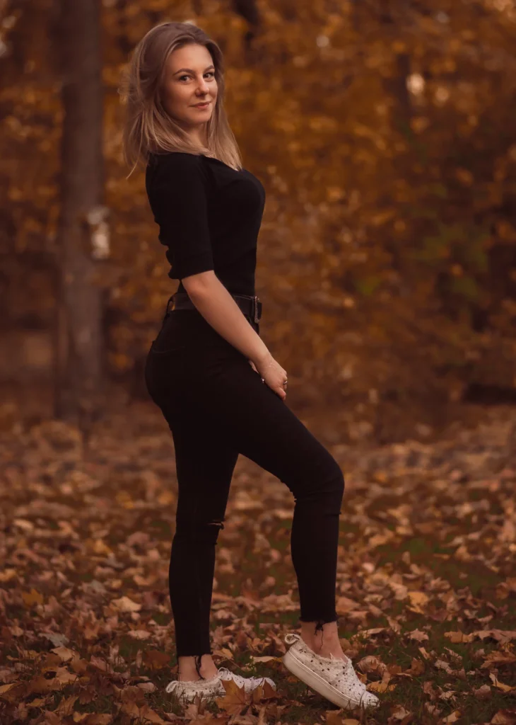 Kobieta w czarnym stroju stojąca w parku pełnym spadających liści jesienią.