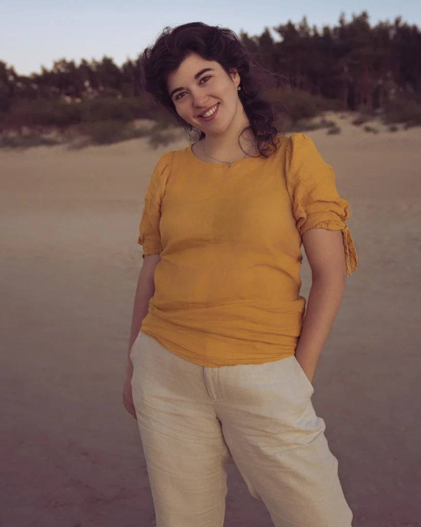 Uśmiechnięta kobieta w żółtej bluzce i lnianych spodniach na plaży.