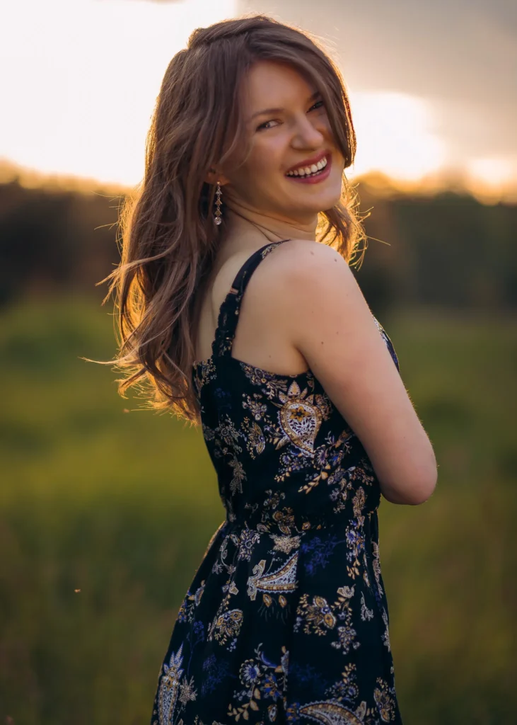 Uśmiechnięta młoda kobieta w sukience z nadrukiem kwiatowym na tle zachodzącego słońca.