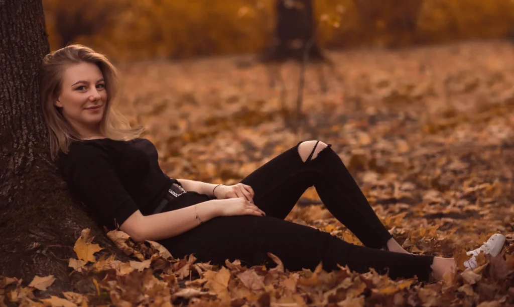 Uśmiechnięta młoda kobieta siedząca na ziemi otoczona przez opadłe liście.