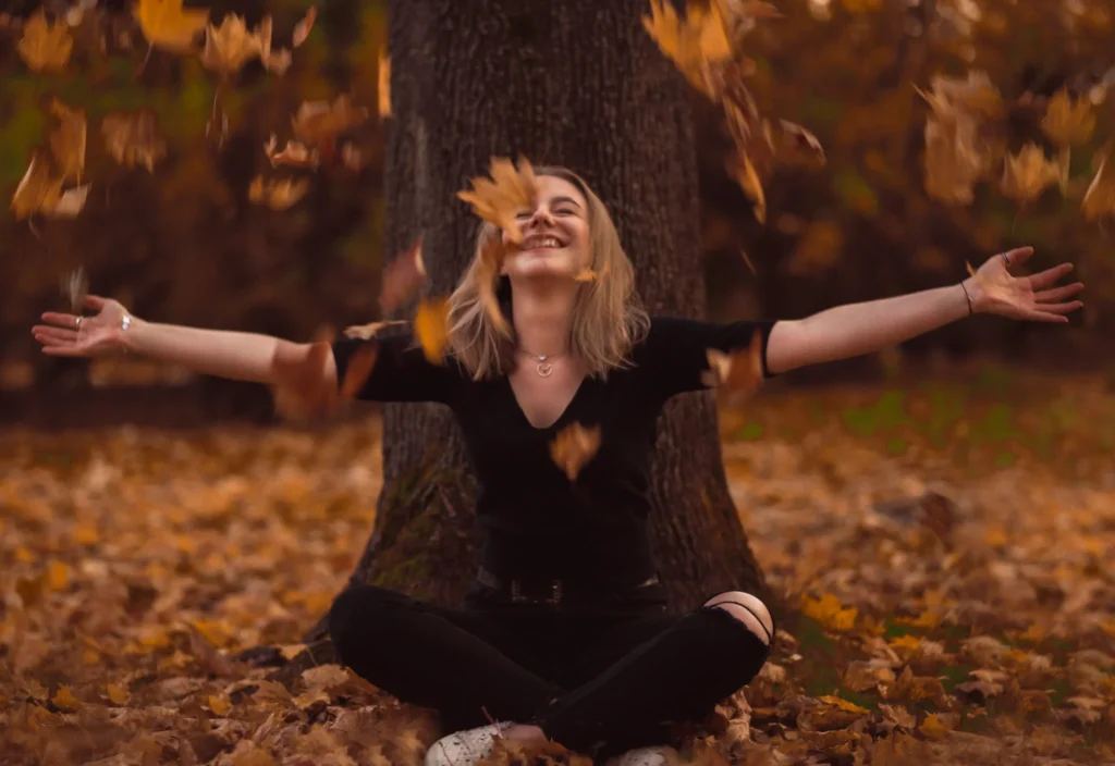 Radosna kobieta siedząca pod drzewem, otoczona przez spadające jesienne liście