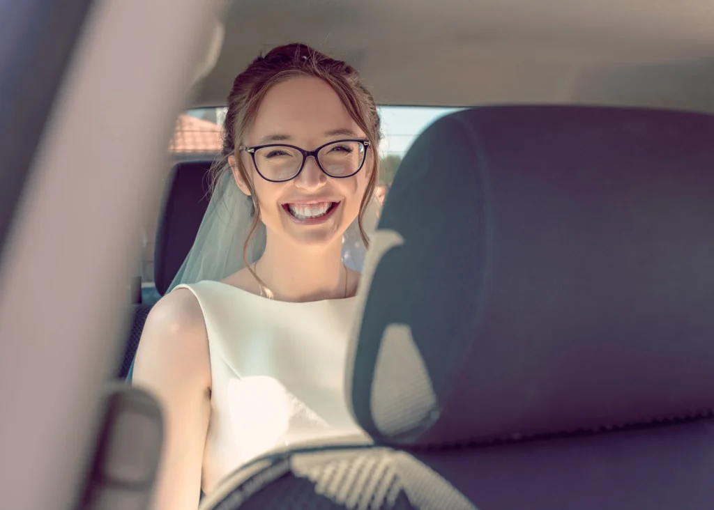 Uśmiechnięta panna młoda w okularach siedzi w samochodzie w drodze na ceremonię ślubną.