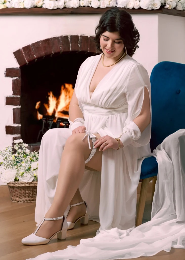 Elegancka fotografia ślubna. Panna młoda w białej sukni ślubnej z perłowym naszyjnikiem uśmiecha się, siedząc przed kominkiem.